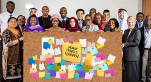 An international group of adults stand with a big board in front of them. It says, Make Things Happen. There are lots of coloured post it notes on the board.