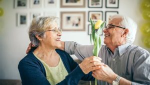 A older man and woman are smiling at each other. The man is handing the woman a yellow tulip. Creating environments for dementia.