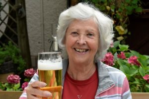 An older woman sits in a garden. She is holding a glass of beer and smiling. She looks happy.