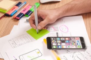 A hand holding a coloured pen is poised over a green post it note. There are drawings on the table and a smartphone. It indicates UX design.  UD, ID, DfA, UX, UA muddle.