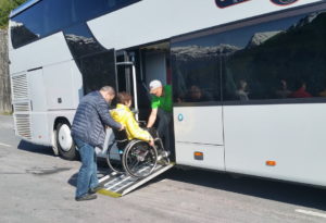 A woman in a yellow jacket is being assisted onto the tour bus by two men up a ramp.