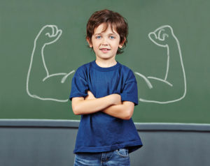 Image of a child with his arms crossed in front of a chalkboard with muscles drawn on it.