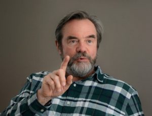 A man in a checked shirt and wearing a beard looks as if he is talking while pointing his finger at someone.