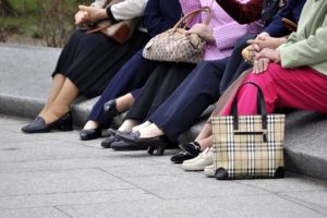 The legs and feet of six older women are shown sitting on a stone wall. They are holding their handbags in their laps. They are wearing sensible shoes.