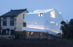 rear view of the all white home at twilight. It shows the ramp coming up from ground level on one side wrapping around the back and up to the first floor on the third side.
