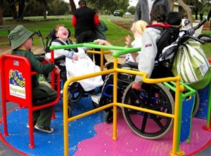 Three children, each a wheelchair user, are enjoying the spinner in the playground: a universal design.