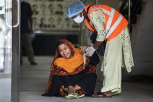 A woman is sitting on the ground and is being helped by a person in protective clothing and a hi vis vest. The woman looks distressed.