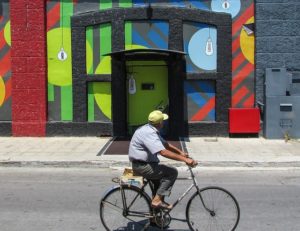 An older man rides his bicycle along a street. In the background is a brightly coloured mural.