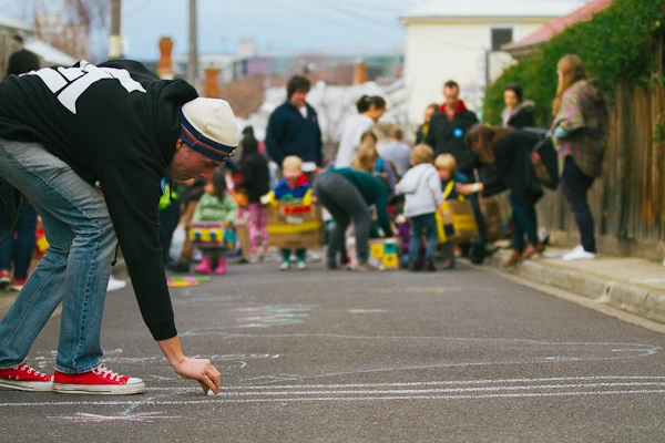 A man is drawing chalk lines on the roadway. In the background children are gathered. 1000 Play Streets Toolkit.