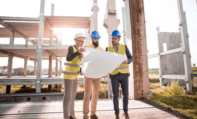 Three men in hard hats stand on a building site looking at architectural design plans.