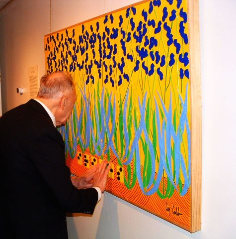 A man in a dark suit places his hands on a brightly coloured painting representing blue flowers on a yellow and orange background. 