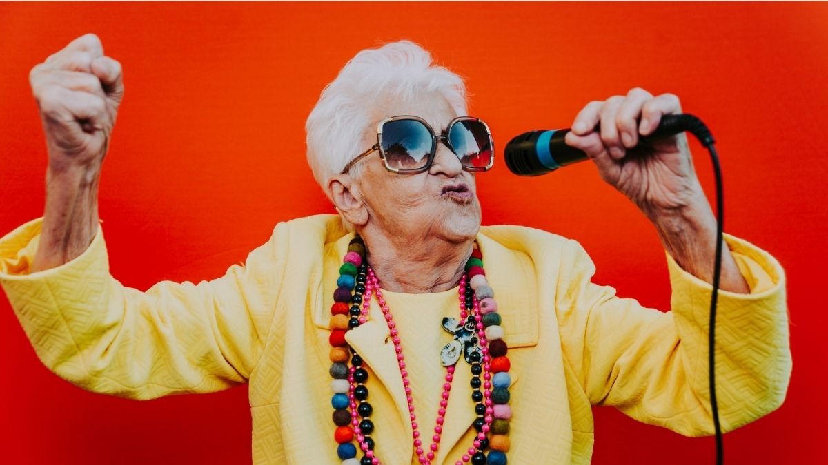 An older woman with white hair and dressed in bright yellow and wearing sunglasses is singing into a microphone enthusiatically on TikTok.