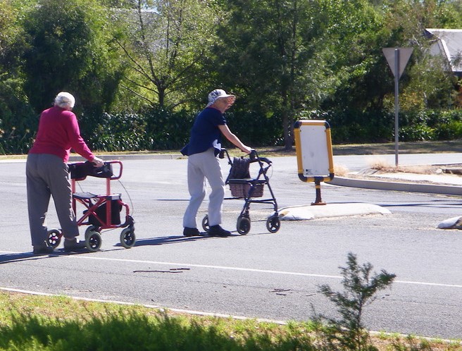 Two women using wheelie walkers are crossing the road in a country town.
