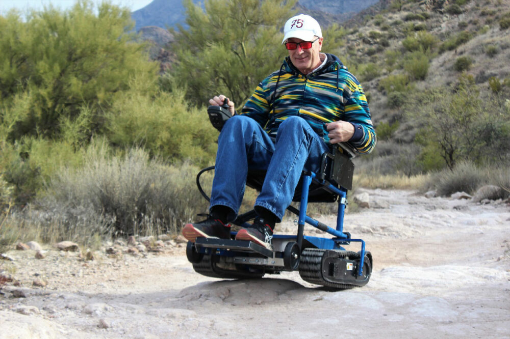 A man in his wheelchair with the Freedom Trax attached. He is on a walking trail. 