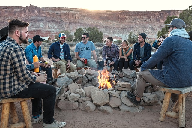 Men are gathered around an outdoor fire in natural surroundings. They are participating in a discussion.