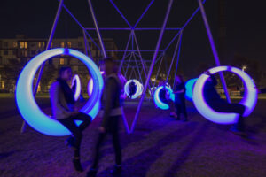 A night time image of a swing set comprising large rings. They are illuminated in purple and blue.