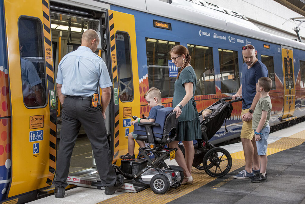 A boy in a powered wheelchair is mounting the ramp into the Queensland Rail train. A woman stands behind him and the station guard looks on. A man with a baby stroller and boy wait nearby to enter the train carriage. The image is from the Access and Inclusion webpage.