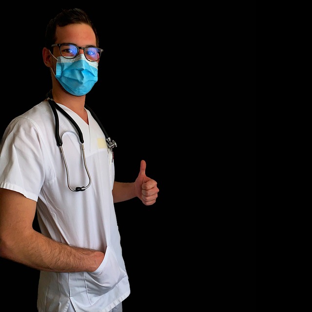 A man in a white hospital shirt is wearing a blue face mask and has a stethoscope around his neck. He is looking into the camera and is posing with a thumbs up sign.